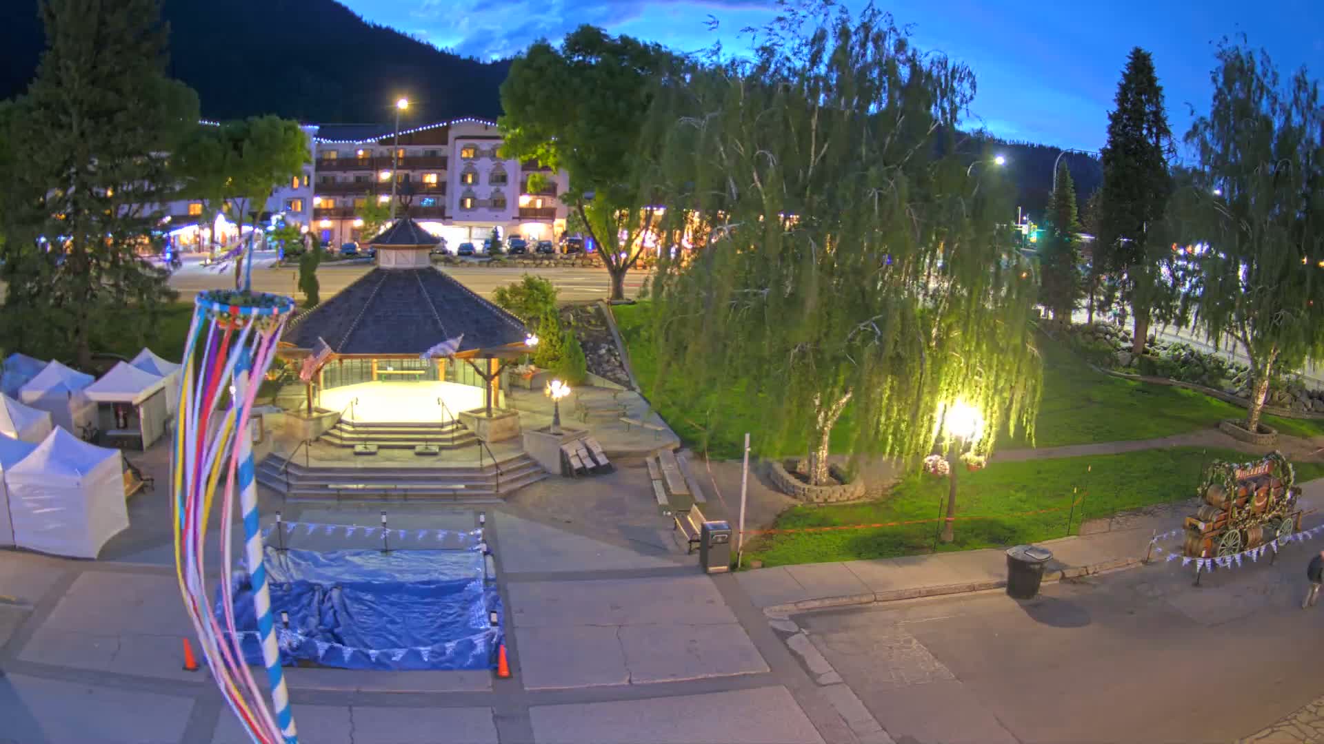 A town square at dusk, featuring a gazebo, a maypole, several tents, and a decorated wagon, is nestled beneath mountains under a twilight sky.