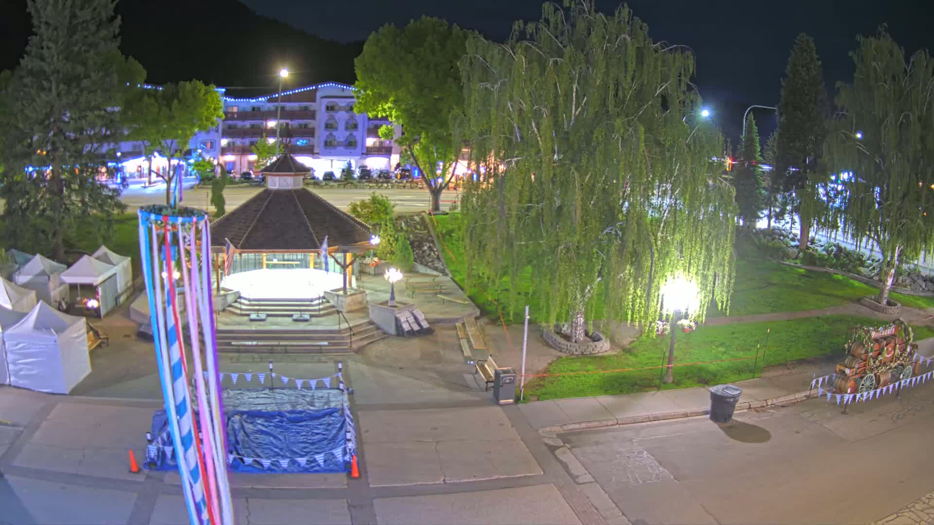 A nighttime view of a town square with a gazebo, several trees, and market stalls, under clear skies.