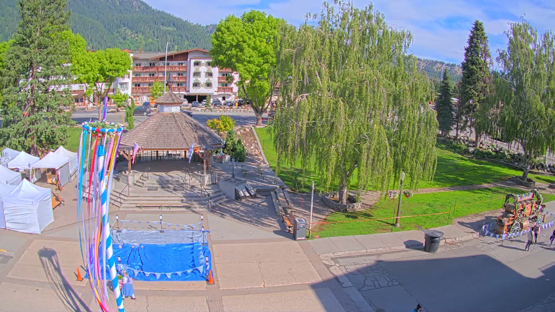 A town square with a gazebo, a maypole, market stalls, and a decorated wagon is situated under a sunny sky, surrounded by trees and a hotel in the background.