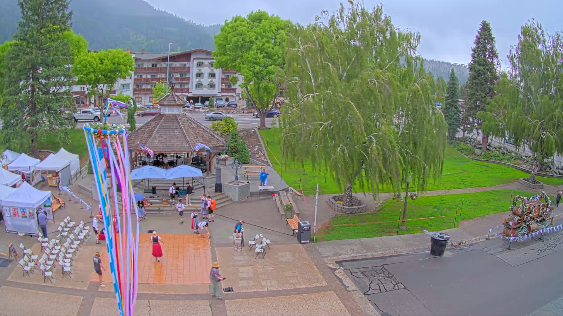 An overcast day reveals a town square with people gathered around a maypole and a dance floor, near a gazebo and vendor tents, with a multi-story building and trees in the background.