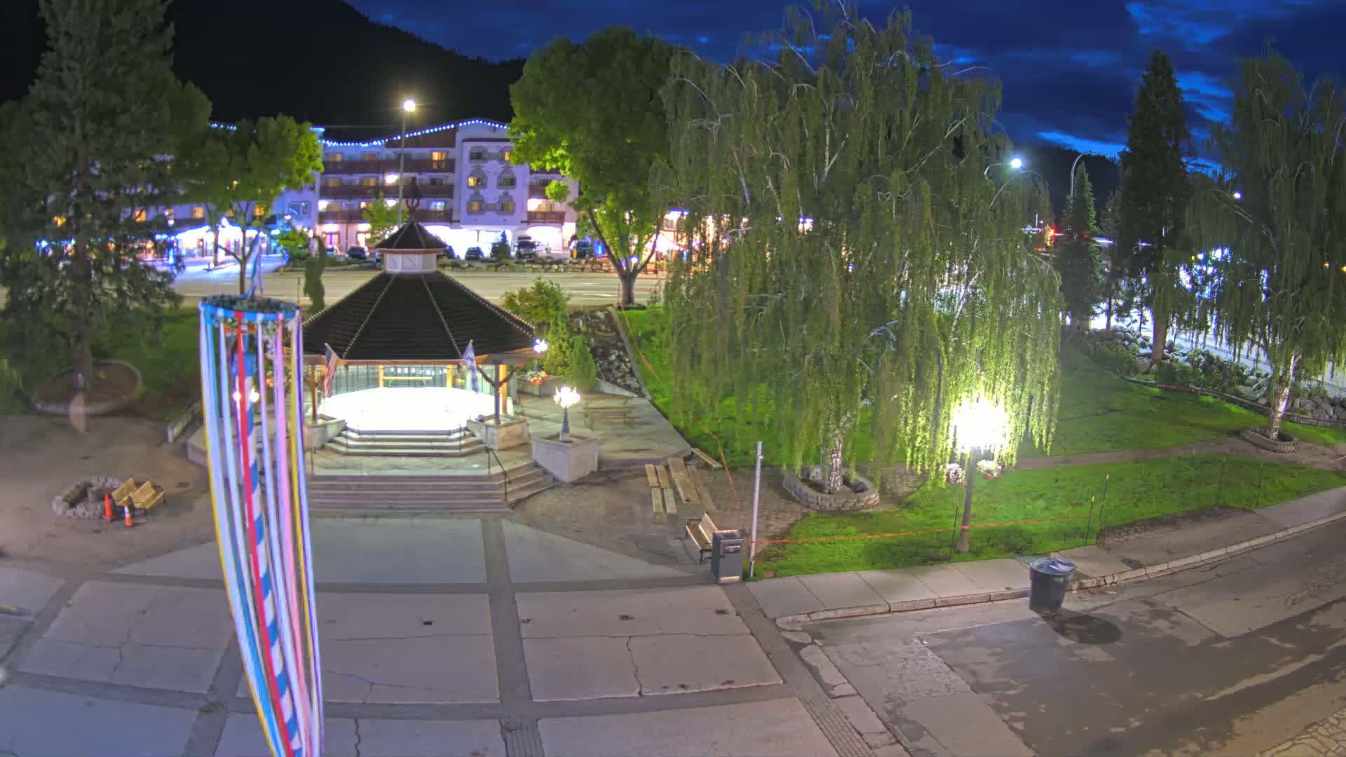 A nighttime view of a town square with a gazebo, trees, and a maypole, under a partly cloudy sky.