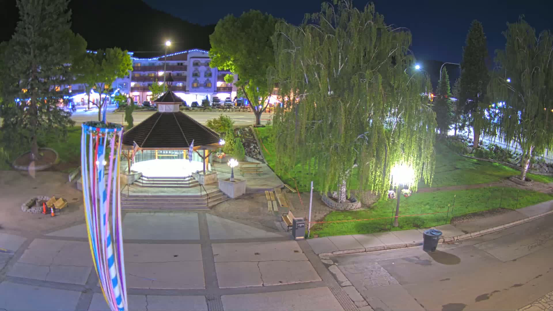A nighttime view of a town square with a gazebo, trees, and a building in the background.