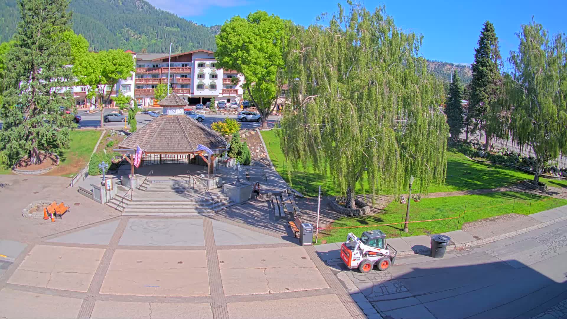 A sunny day reveals a town square with a gazebo, a small park, a multi-story building, and a small construction vehicle.