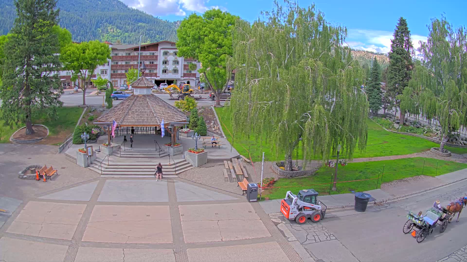 A town square with a gazebo, benches, and landscaping under a sunny sky, with a horse-drawn carriage and small construction equipment nearby, nestled against a backdrop of mountains.