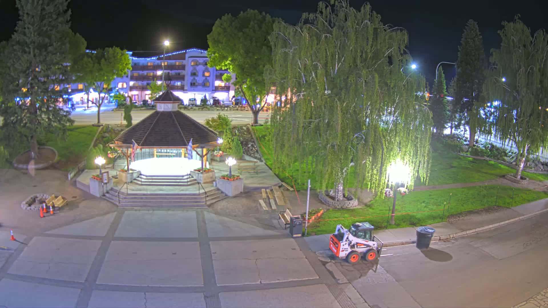 A nighttime view of a town square featuring a gazebo, landscaping, streetlights, and a small construction vehicle.