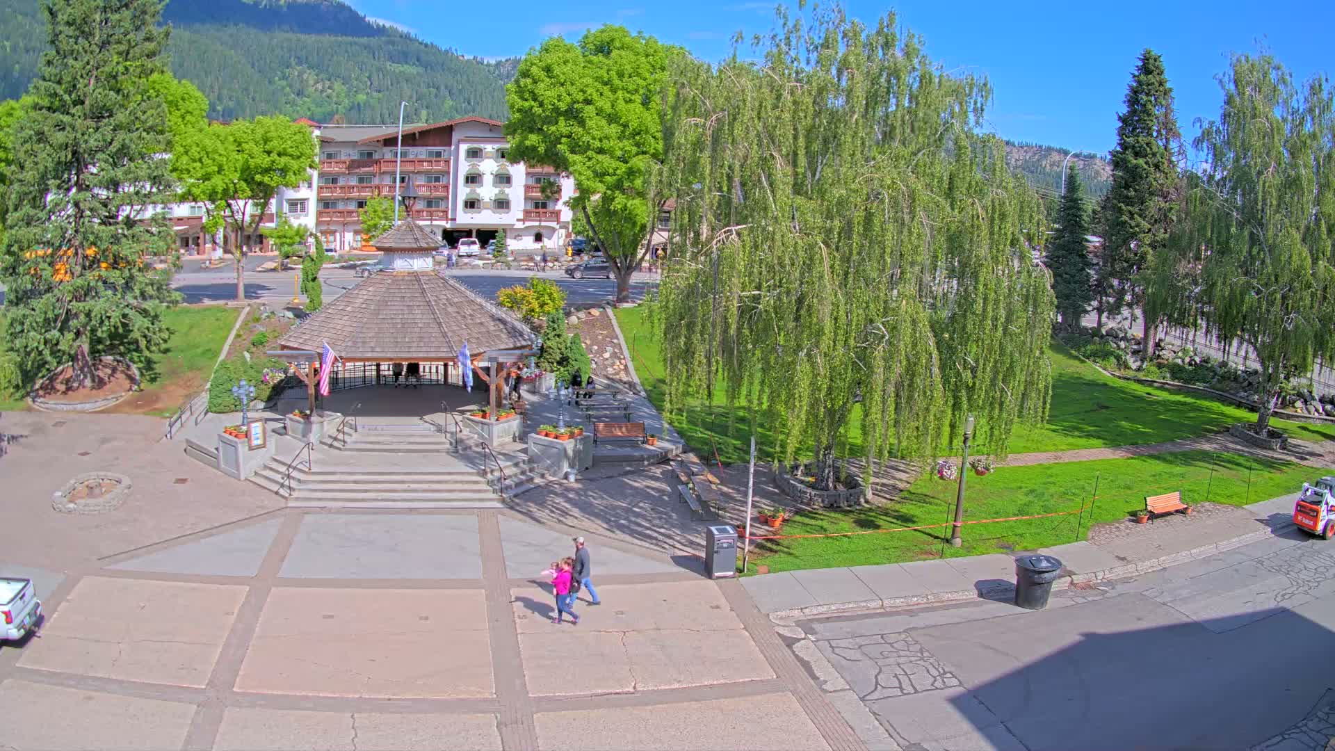 A paved town square with a gazebo, grassy areas, trees, and a multi-story building in the background, under sunny skies.