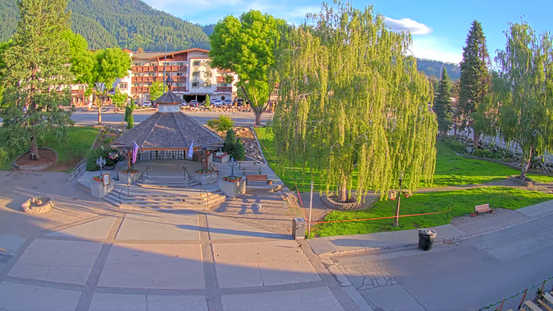 A gazebo sits in a sunny town square, surrounded by trees and a multi-story building in the background.