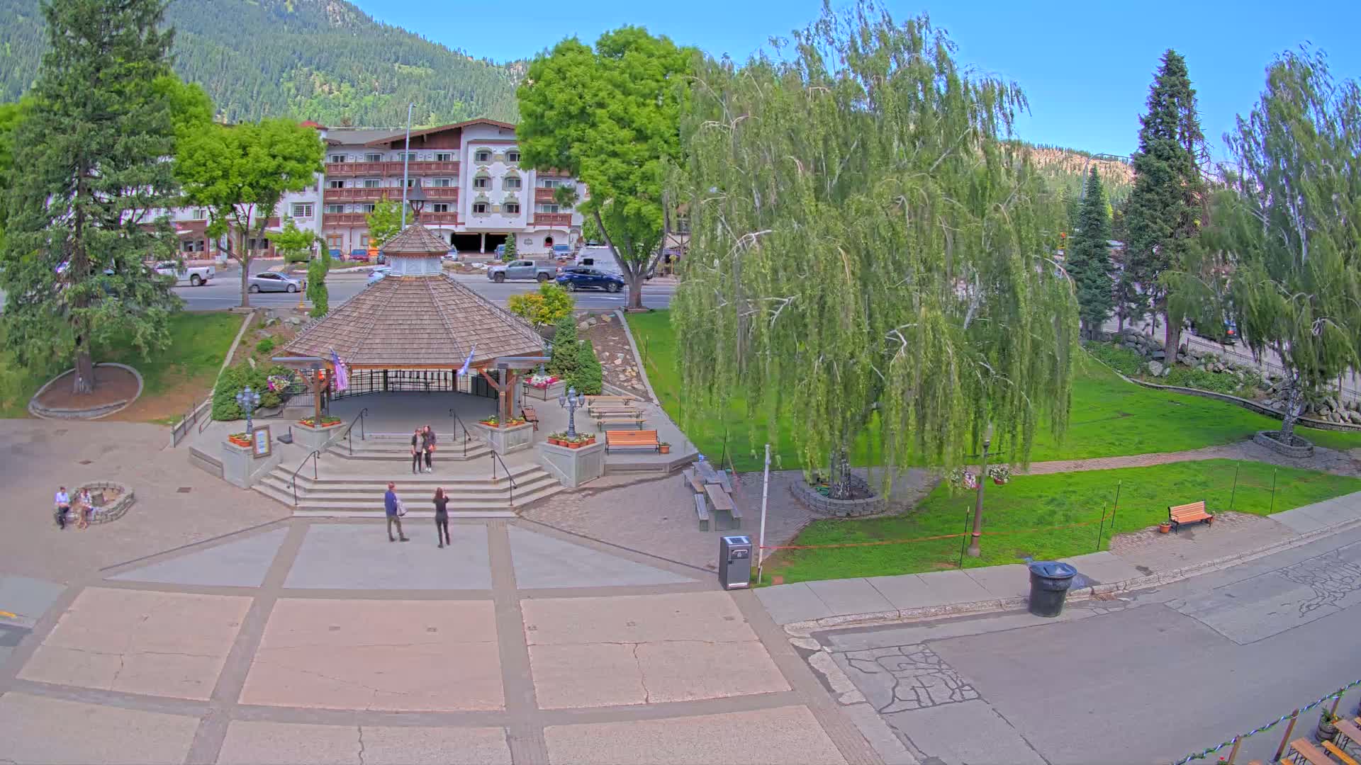 An aerial view shows a town square with a gazebo, several people, lush green lawns, and trees, under a sunny sky.