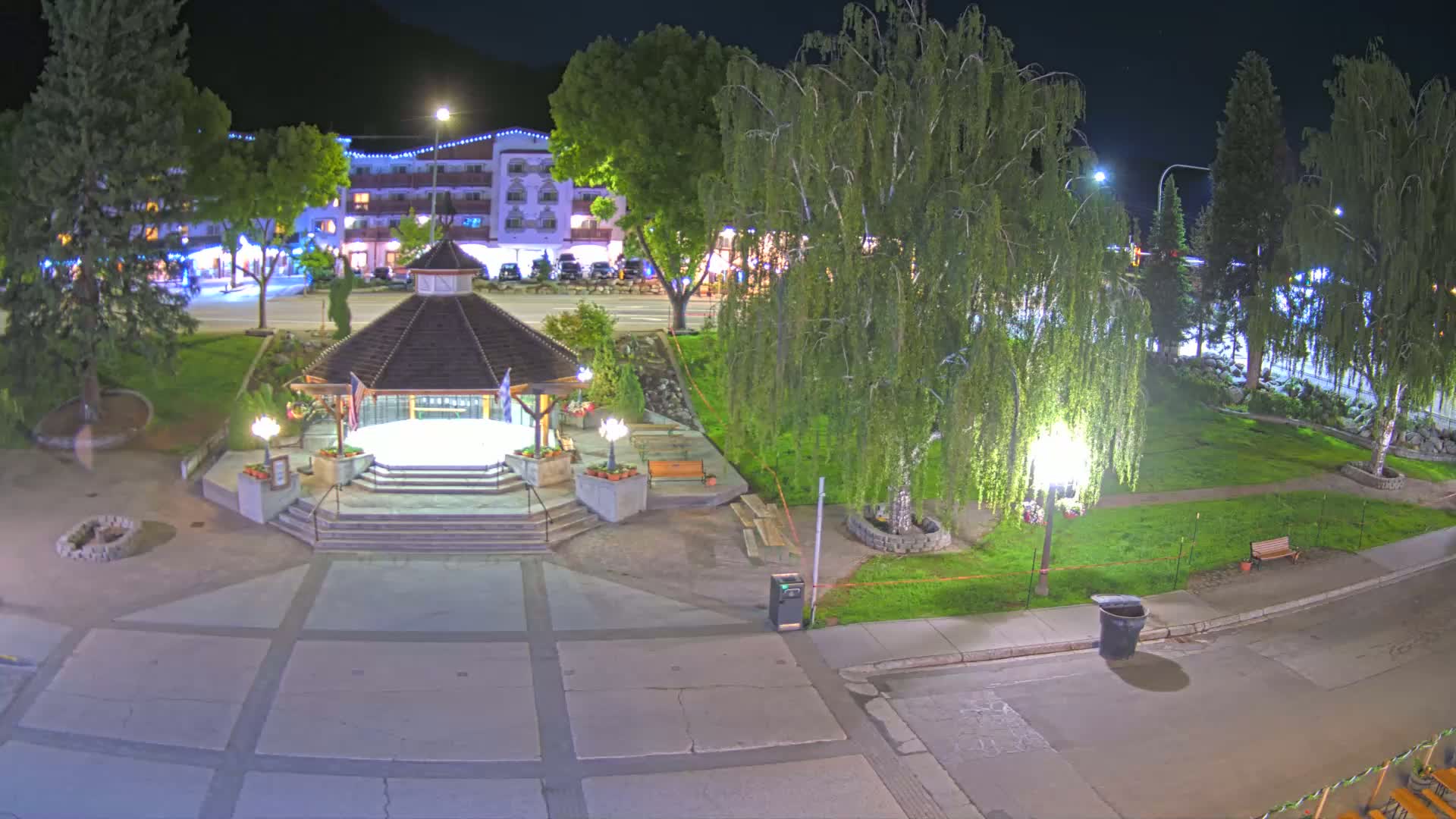 A nighttime view of a town square with a gazebo at its center, surrounded by trees, grass, and a building in the background.