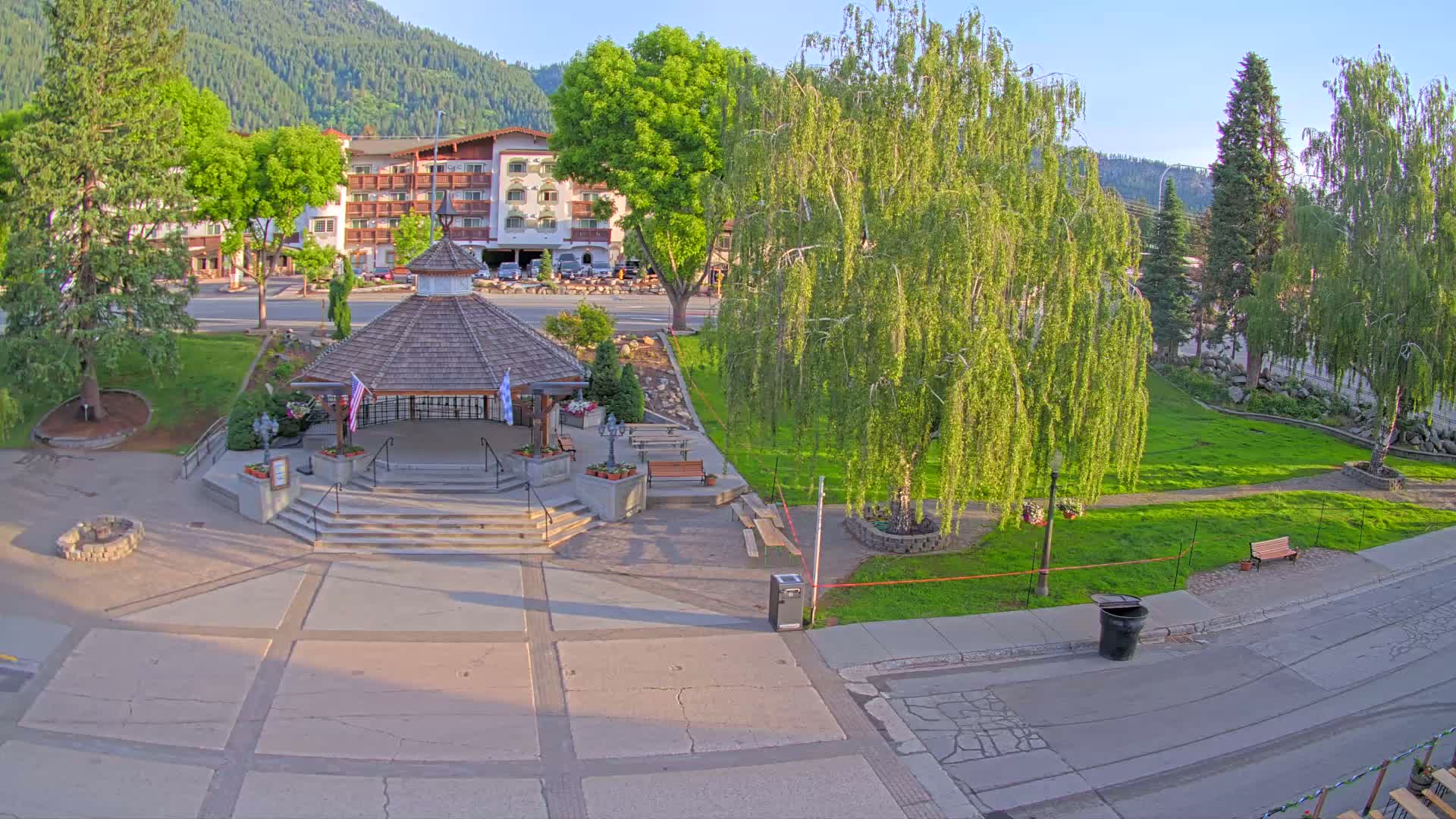A gazebo sits in a town square surrounded by lush green grass, trees, and a multi-story building, under a sunny sky.
