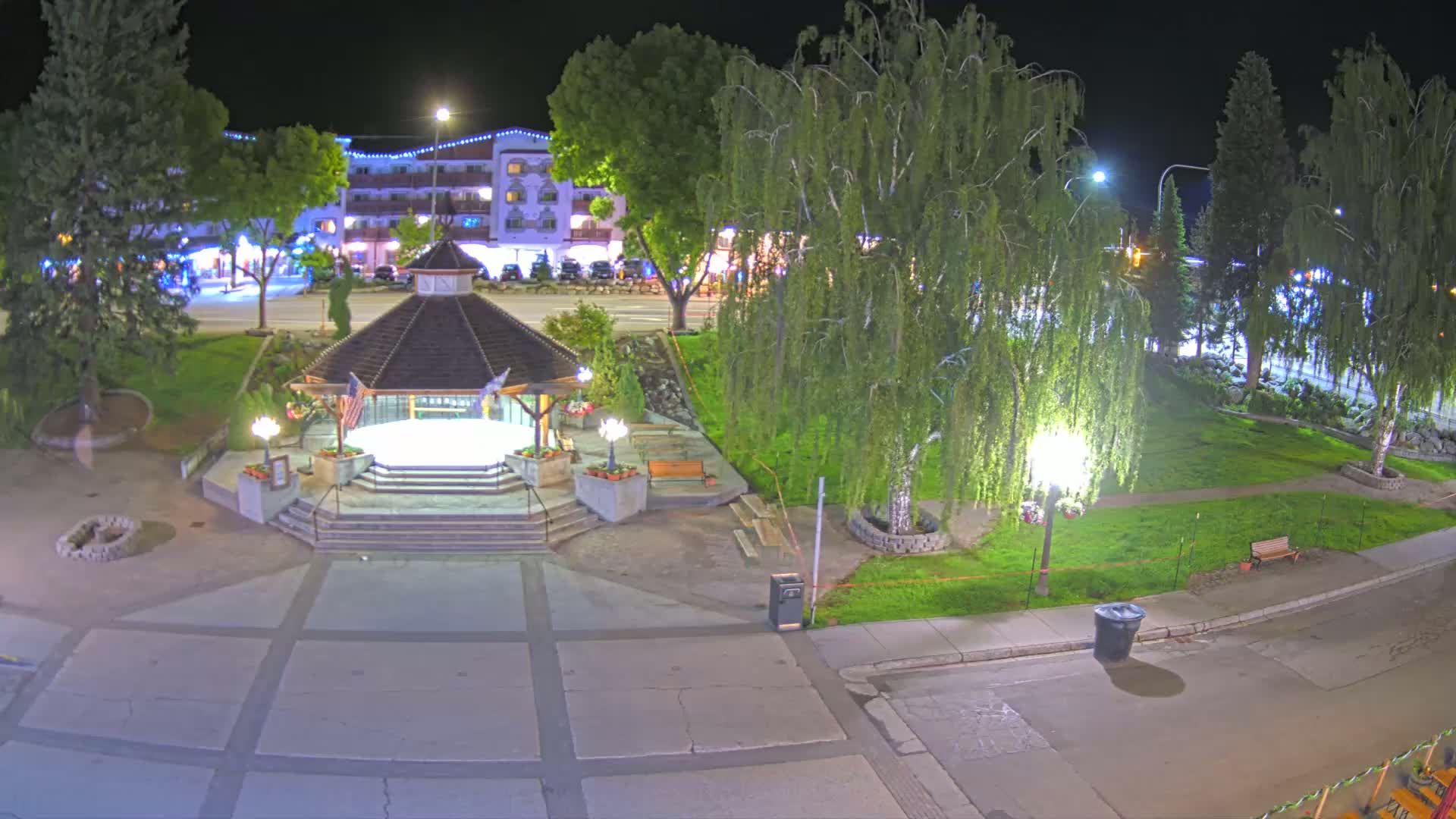 A nighttime view of a town square featuring a gazebo, surrounding landscaping, and a building in the background under clear skies.