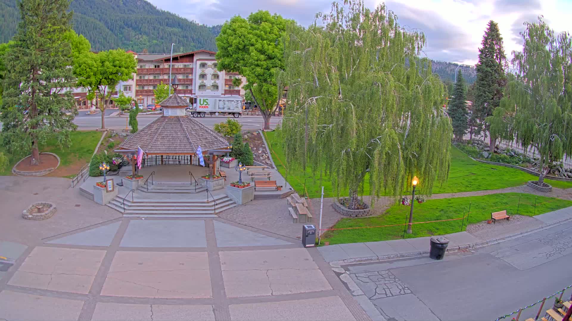 An elevated view shows a gazebo in a park, with a hotel and mountains in the background, under partly cloudy evening skies.