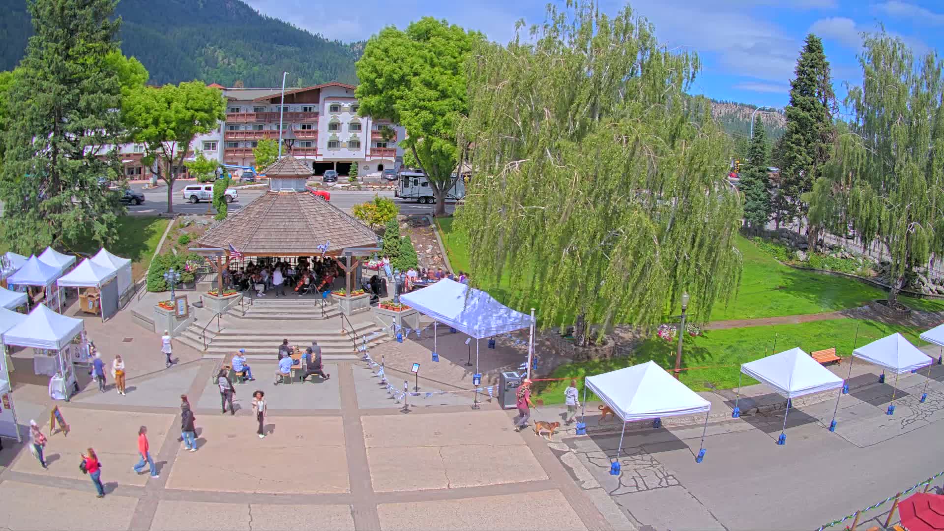 A town square with a gazebo, several white tents, and people milling about on a sunny day, with green grass and trees surrounding it, backed by a mountain range.