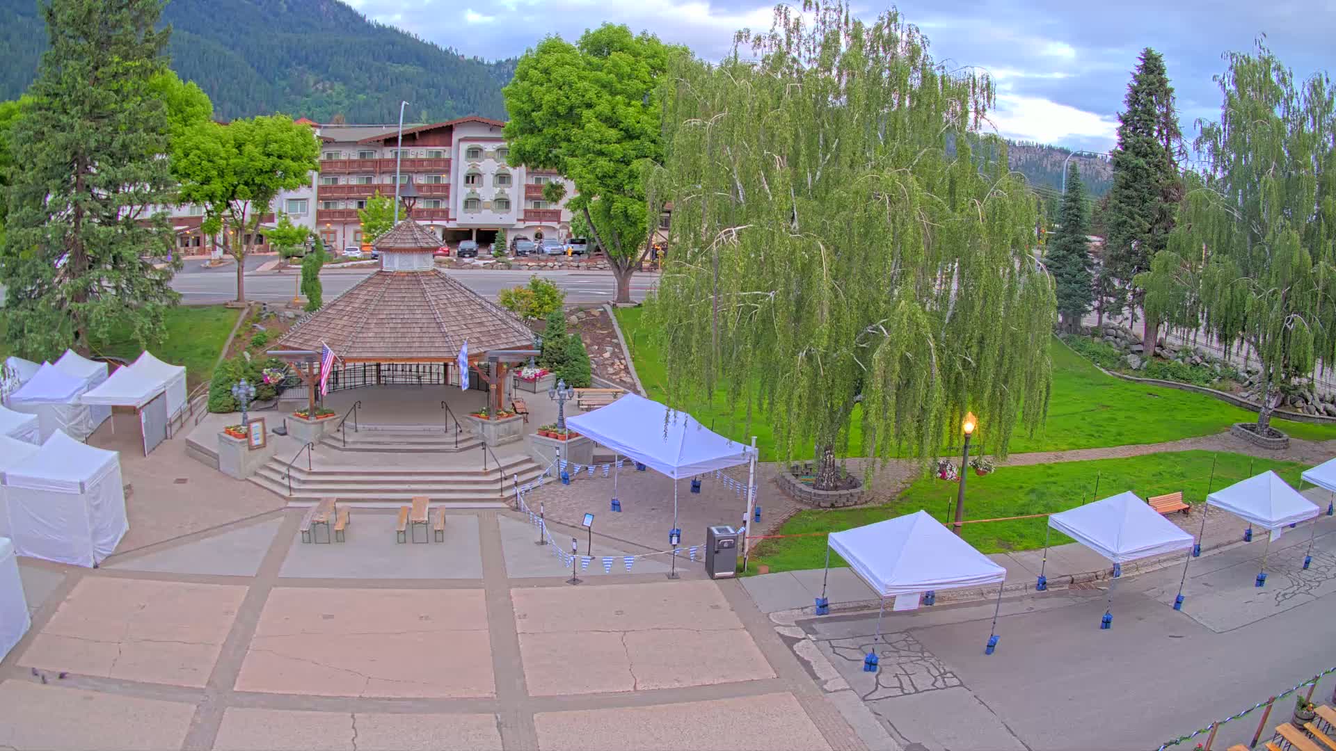 An outdoor town square with a gazebo, several white tents, and a large, lush green lawn under a partly cloudy sky.