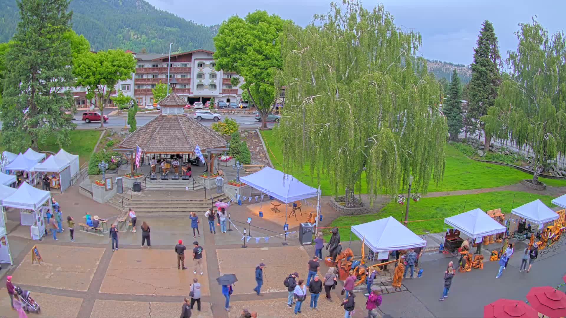 An outdoor arts and crafts fair is taking place in a town square on a cloudy day, with a band playing in a gazebo and numerous people browsing the vendor booths.