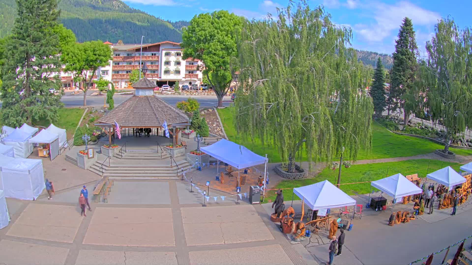 A town square with a gazebo, several white tents displaying crafts, and a hotel in the background on a sunny day.