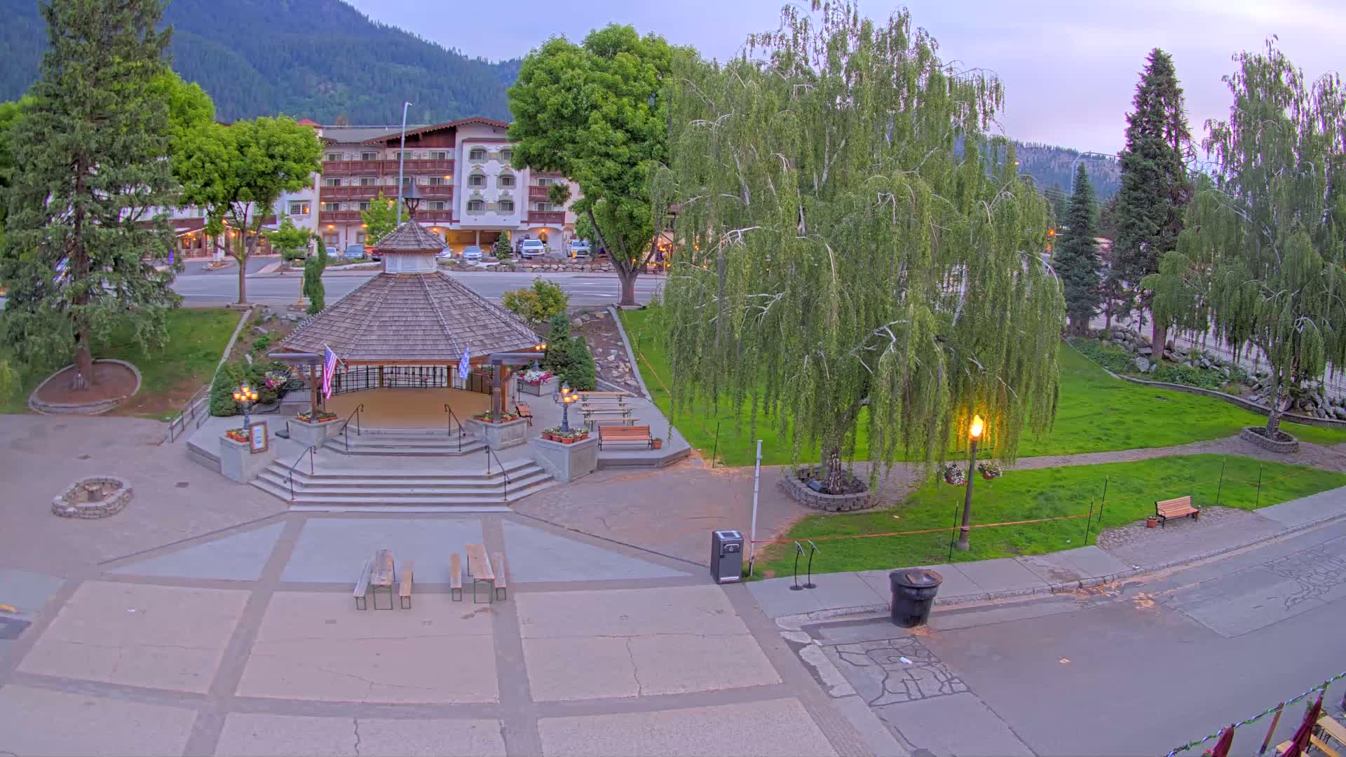 An elevated view of a town square at dusk featuring a gazebo, benches, trees, and a hotel in the background.