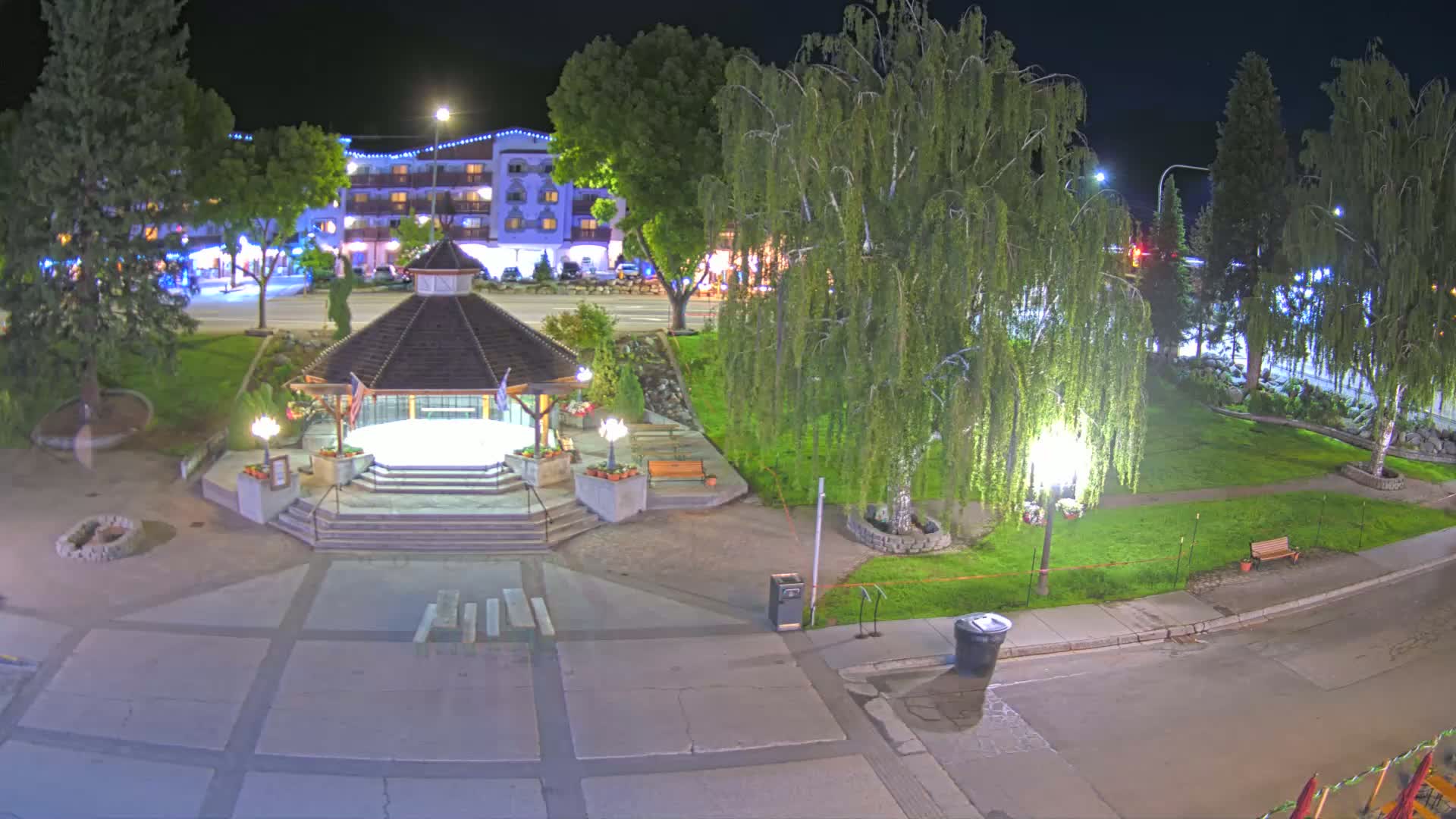A nighttime aerial view shows a gazebo in a town square, with a multi-story building in the background under dark skies.
