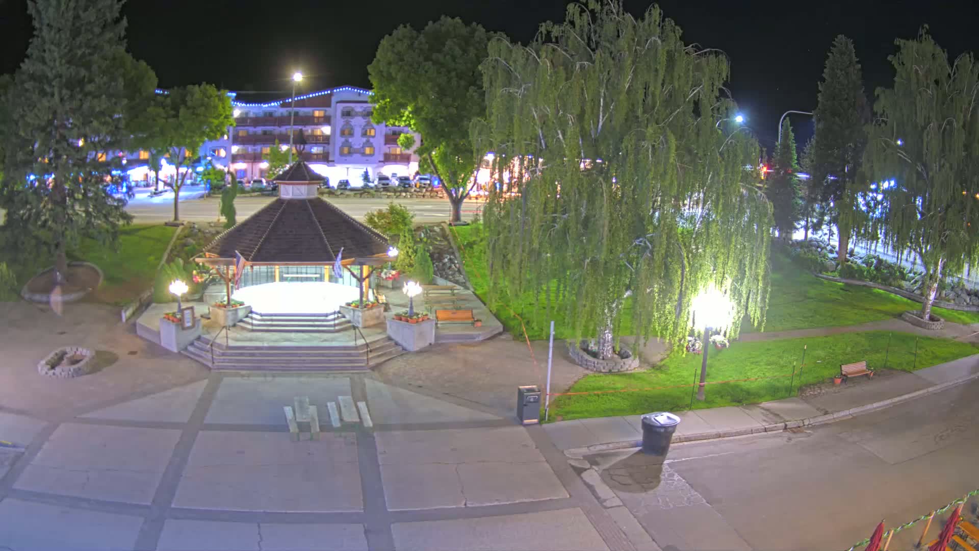 A nighttime view of a gazebo in a park, illuminated by streetlights, with a multi-story building visible in the background.