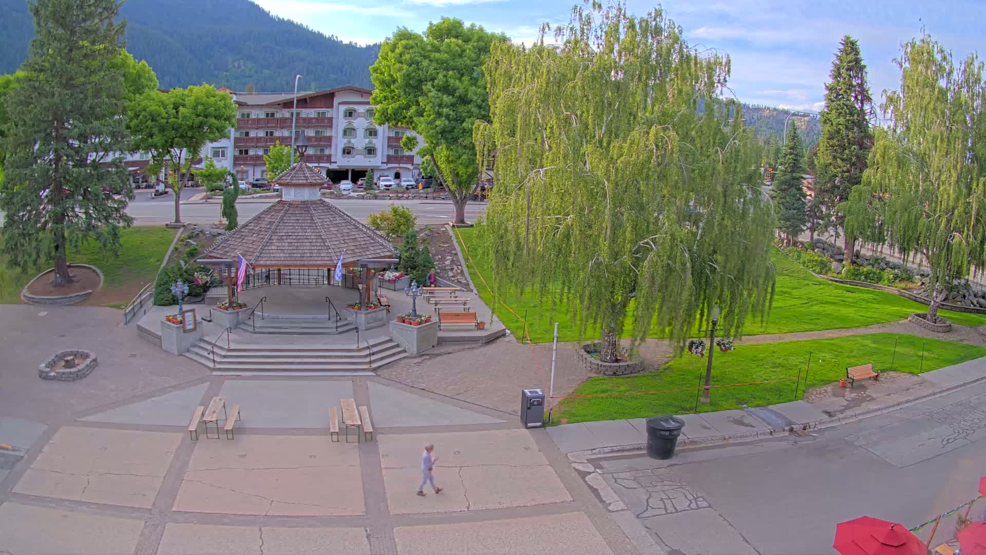 A gazebo sits in a grassy town square under a partly cloudy sky, with a person walking nearby and a hotel visible in the background.