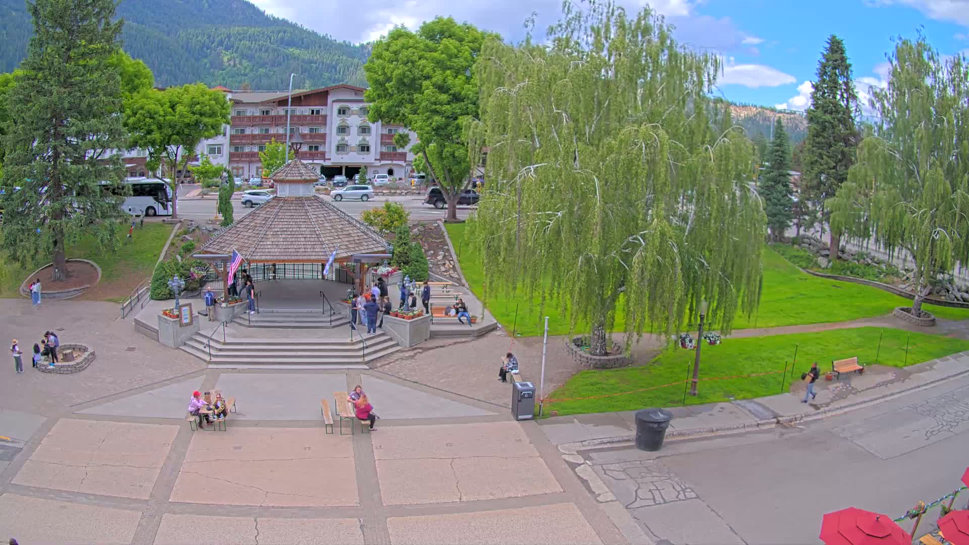 A town square with a gazebo, people sitting at picnic tables, and lush green lawns under a partly cloudy sky, with mountains in the background.