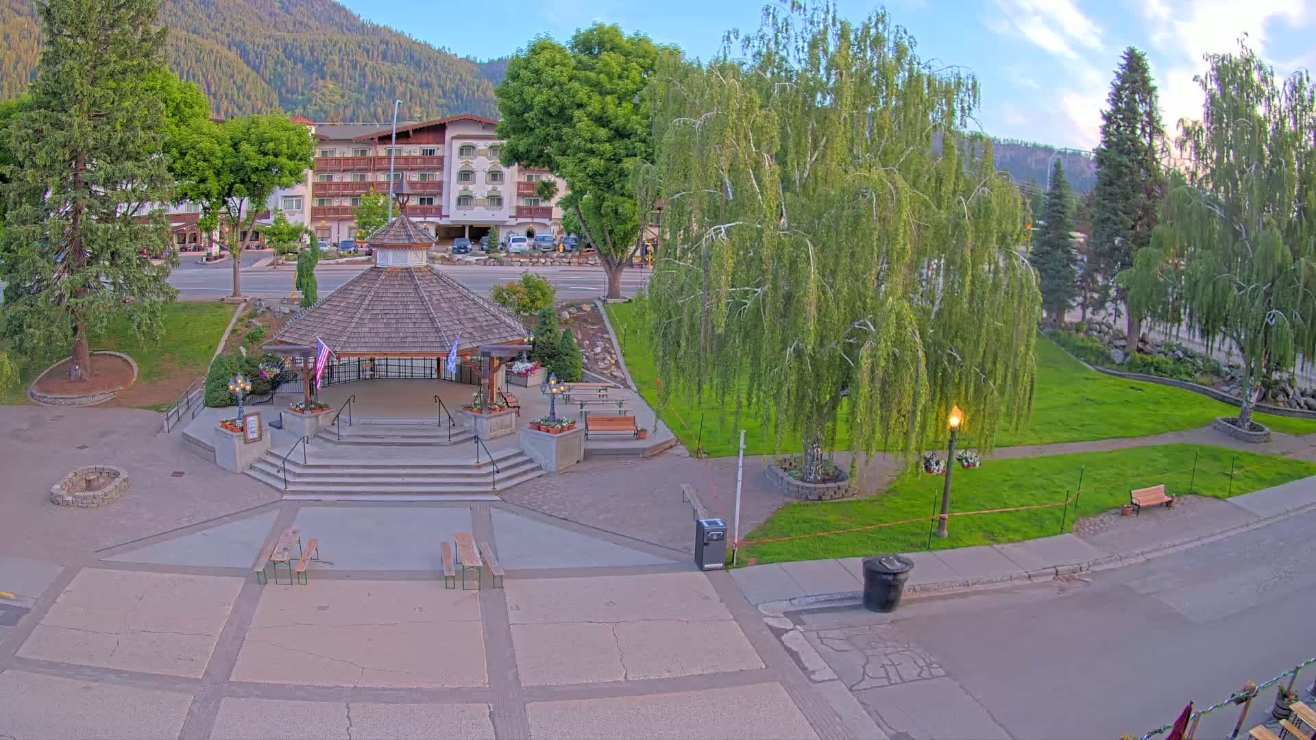 A gazebo sits in a town square next to a grassy area with trees and a hotel in the background, under a partly cloudy evening sky.