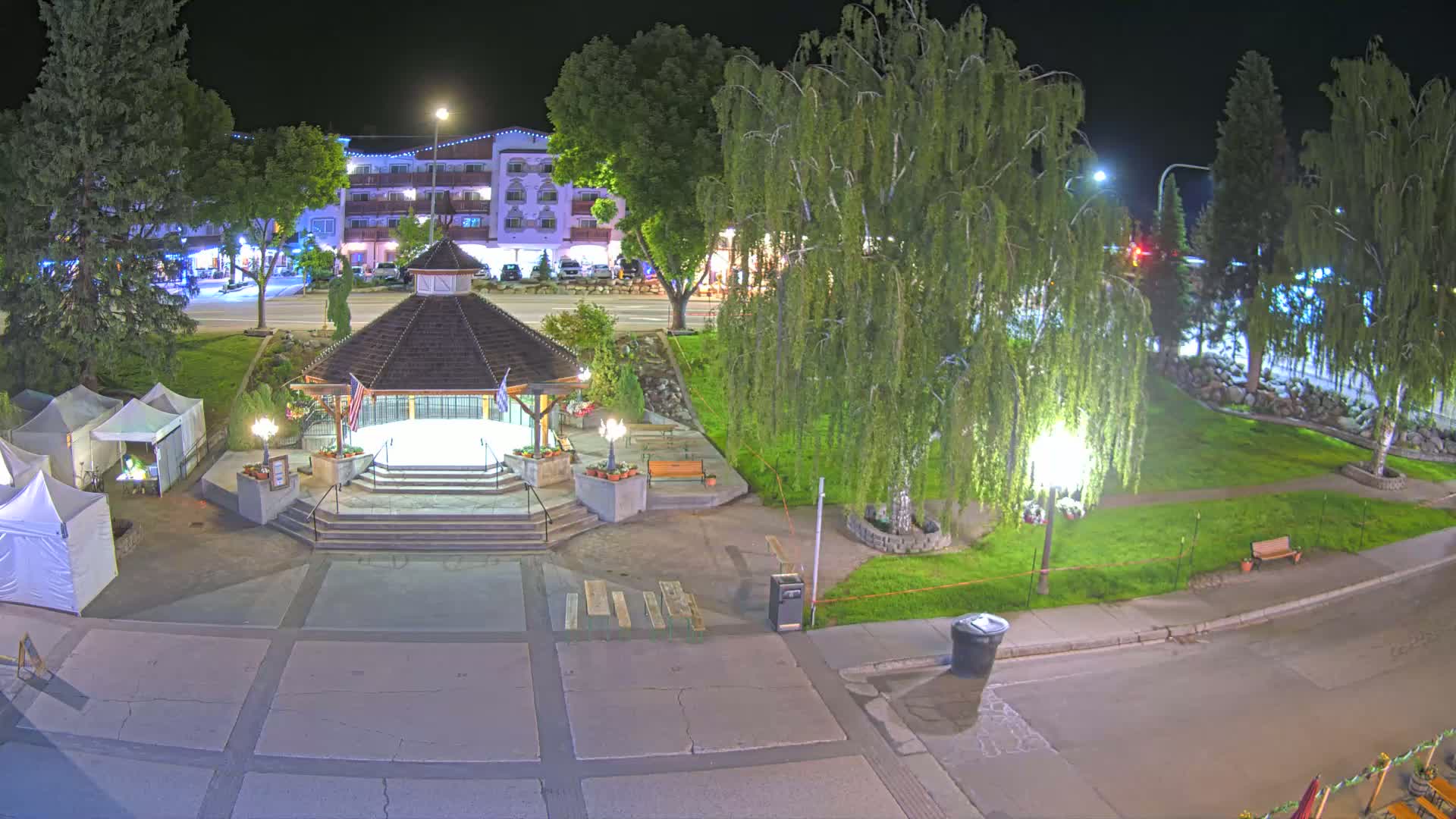 A nighttime view of a town square with a gazebo, surrounded by trees, grass, and a few small tents, under clear skies.