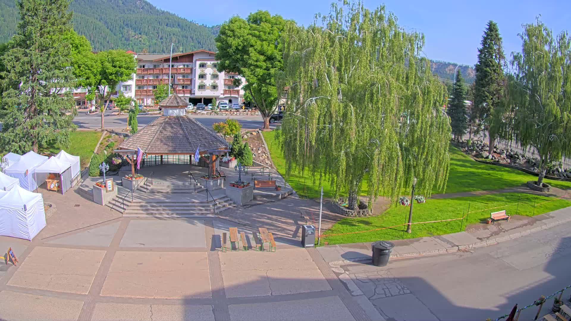An elevated view of a town square on a sunny day shows a gazebo, several picnic tables, some small tents, and a grassy area with trees, all situated near a multi-story building and mountains in the distance.