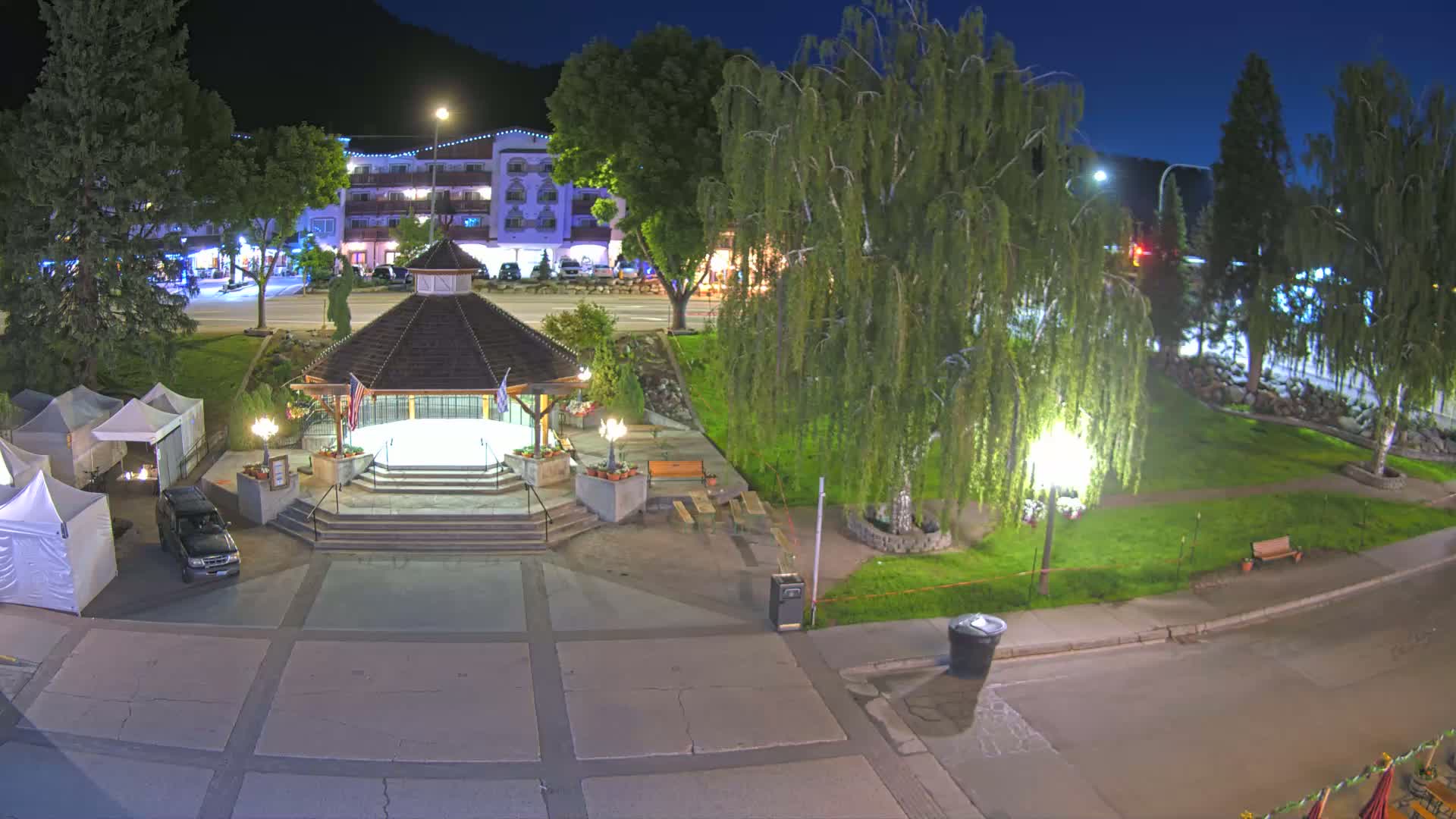 A nighttime view of a town square featuring a gazebo, a few parked cars, and surrounding buildings under a clear, dark sky.