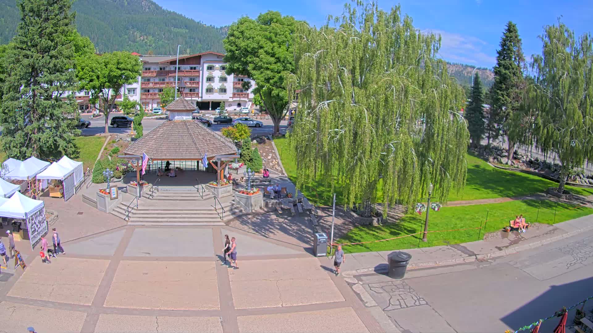 A town square with a gazebo, several people, some market stalls, and lush green trees under a sunny sky.
