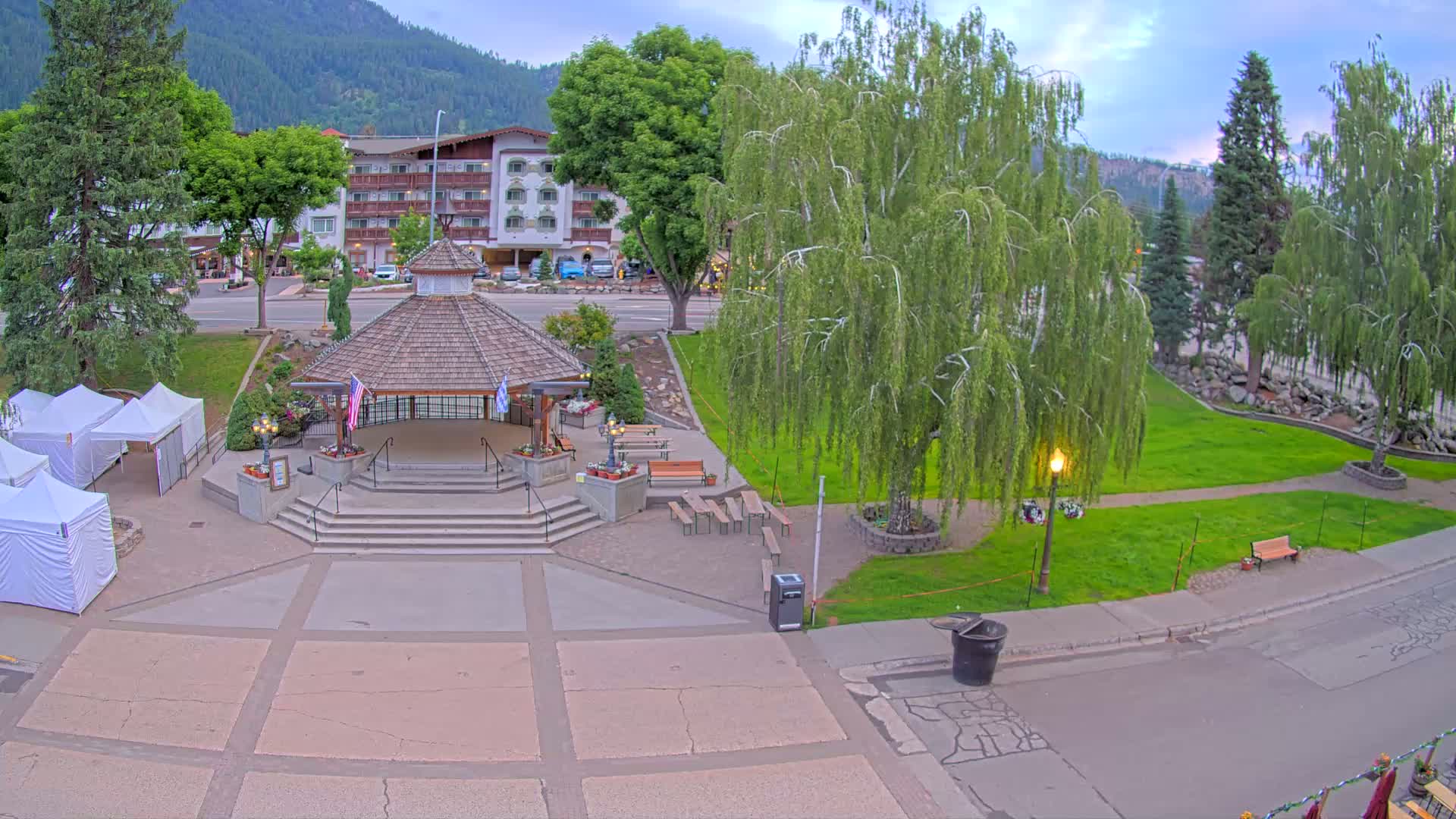 A gazebo with stairs sits in a town square surrounded by trees, picnic tables, and white tents under a partly cloudy sky.