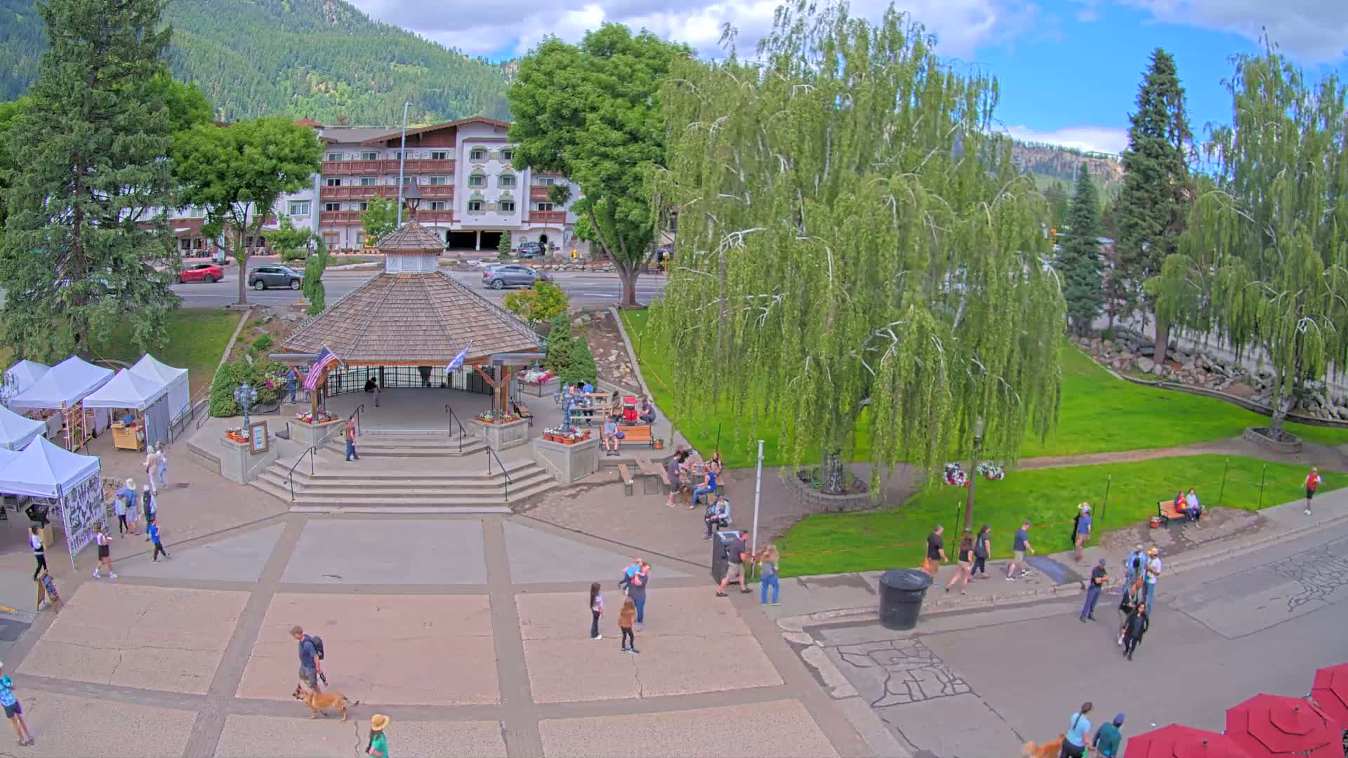 A town square with a gazebo, surrounded by people, green grass, trees, and a hotel, under partly cloudy skies.