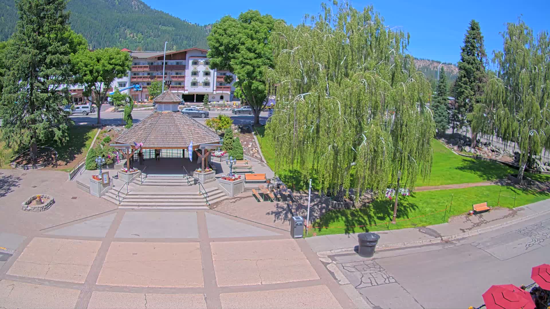 A park with a gazebo and several trees sits in front of a multi-story building under a sunny sky.