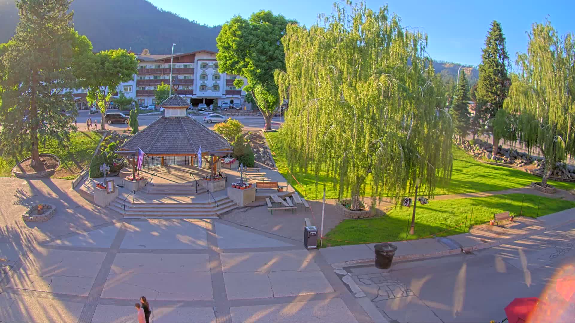 A gazebo sits in a park with a grassy area, trees, and a building in the background, all under sunny skies.