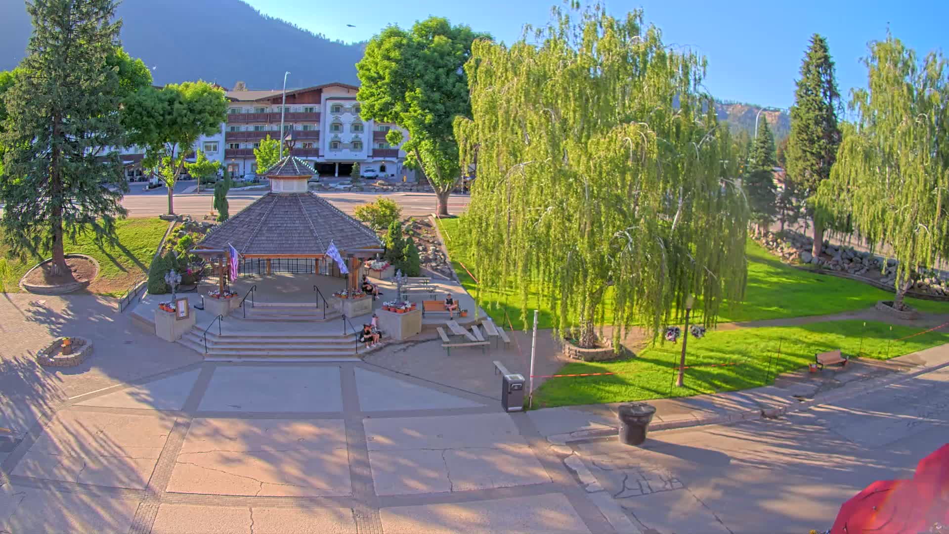 A gazebo sits in a park with several trees, a hotel in the background, and under sunny skies.