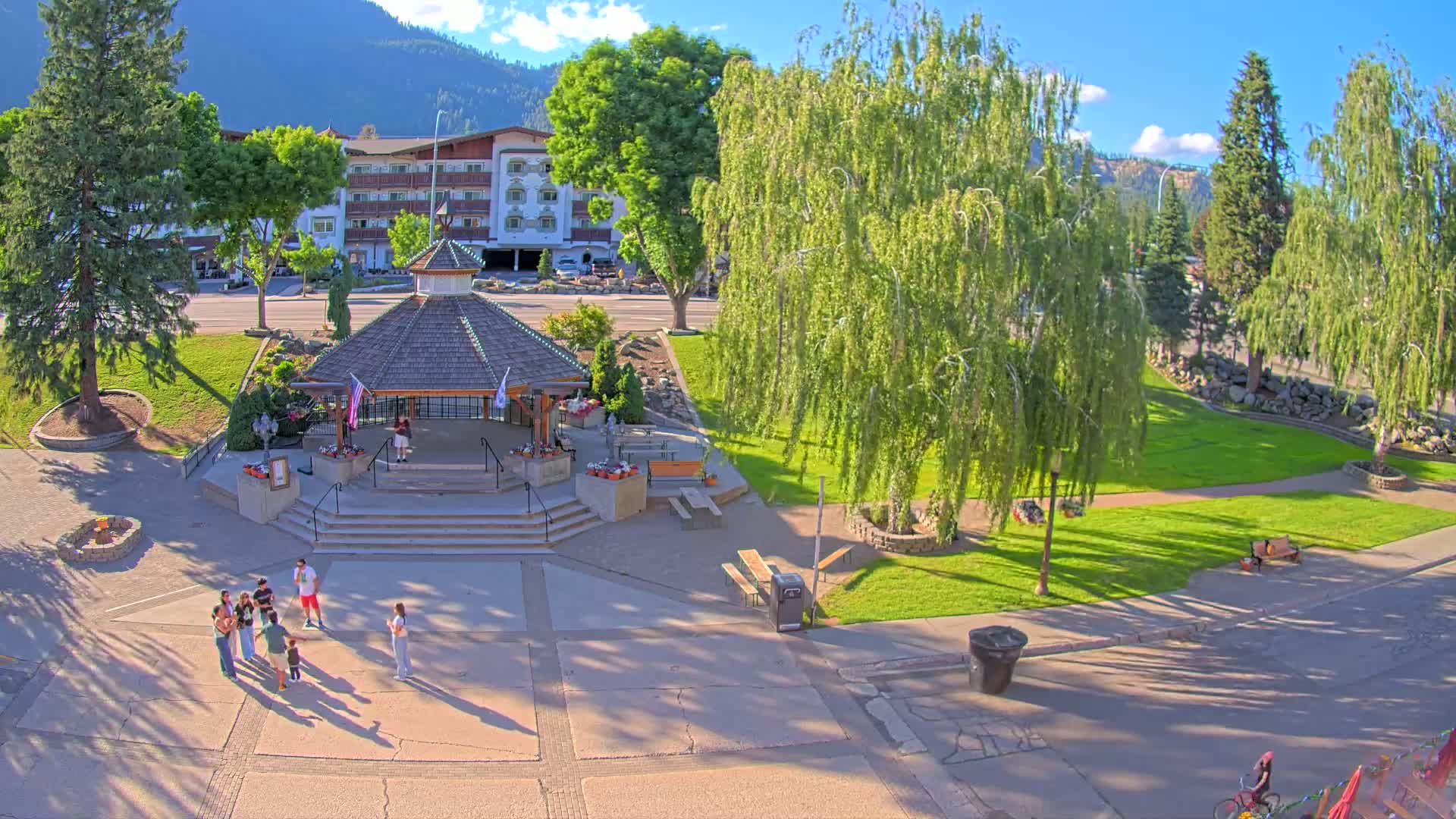 A park with a gazebo, several trees, and people standing in a plaza on a sunny day, with mountains in the background.