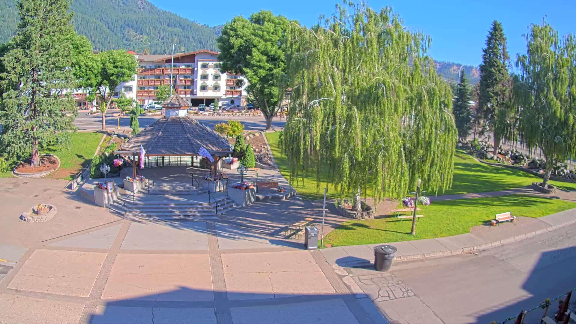 A park with a gazebo and a hotel in the background on a sunny day, surrounded by trees and landscaping.