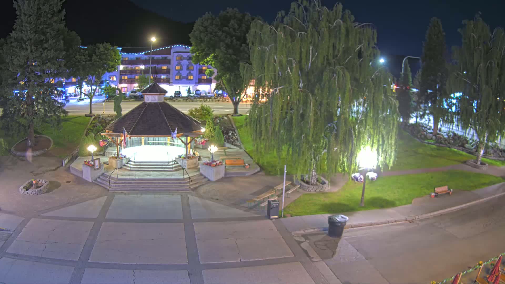 A nighttime view of a park with a gazebo, some trees, and a building across the street, under clear skies.