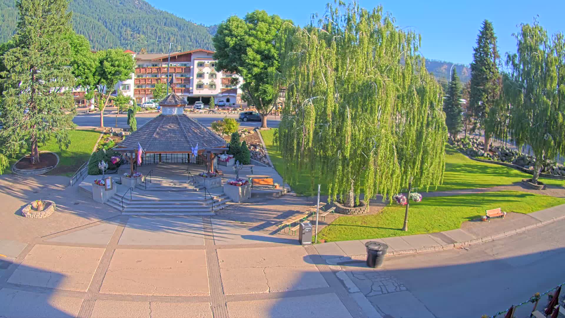 A park with a gazebo and several trees sits in front of a building on a sunny day in a mountain valley.