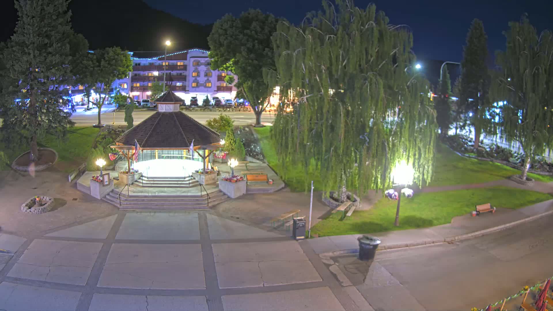A nighttime view of a town square with a gazebo, trees, and a multi-story building in the background under clear skies.