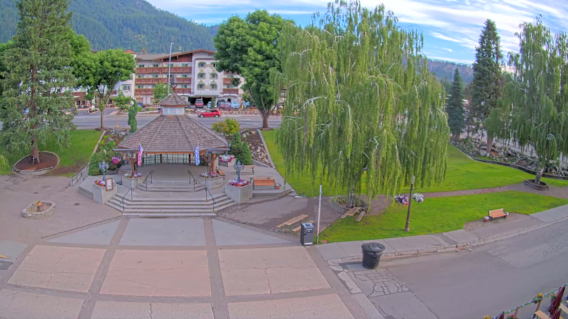 A gazebo sits in a town square with grassy areas, trees, and a hotel in the background on a partly cloudy day.