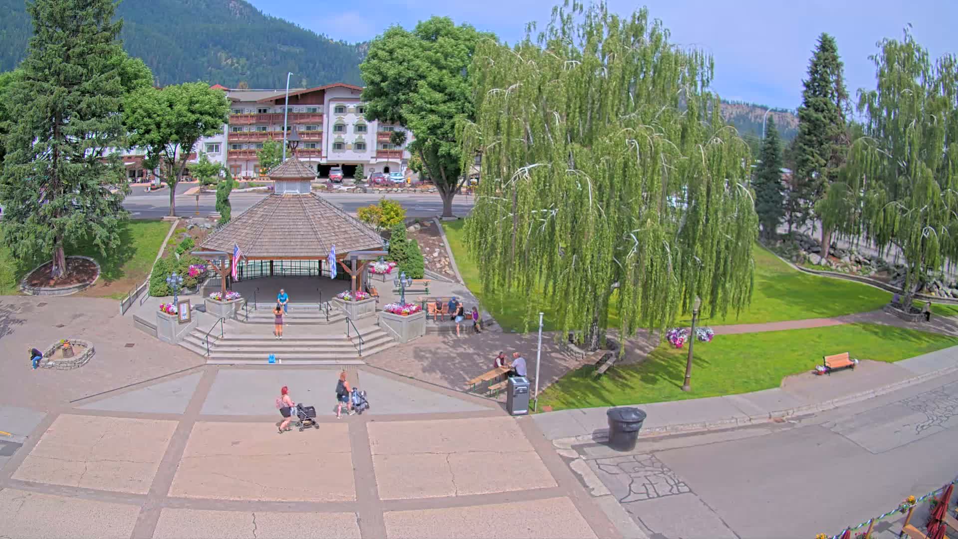 A park with a gazebo, several people, and a hotel in the background on a sunny day.
