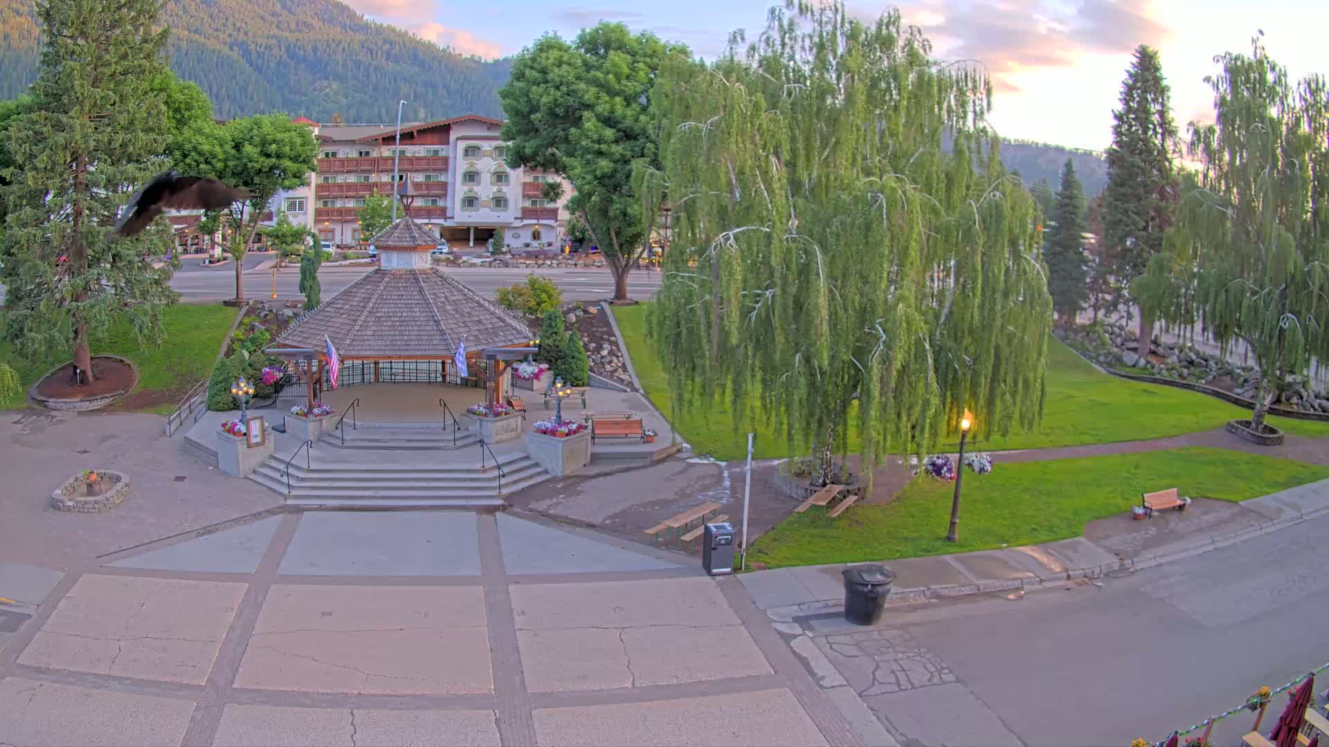 A bird flies over a town square containing a gazebo, several trees, benches, and a paved area, all under a partly cloudy evening sky.