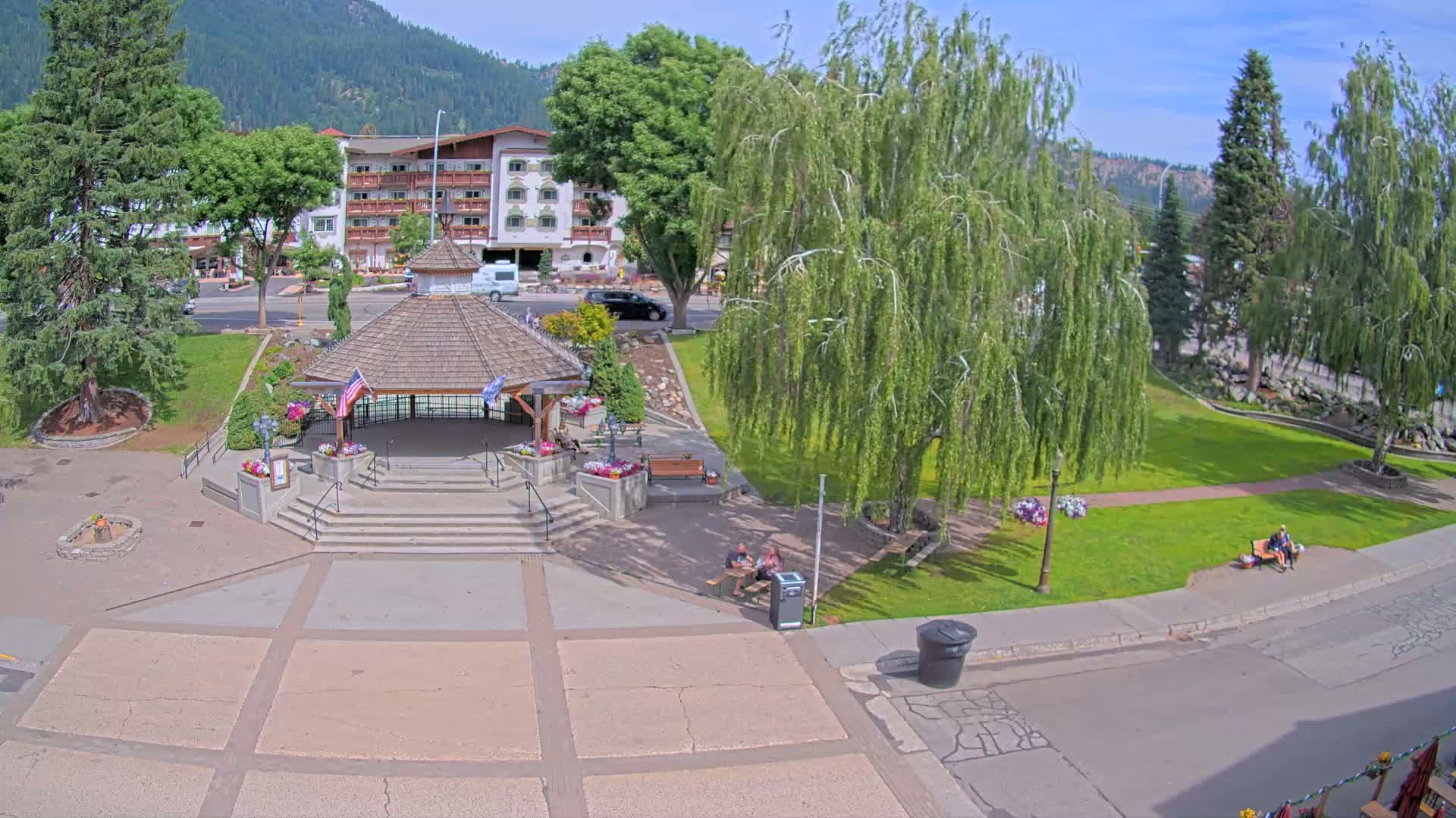 A paved town square with a wooden gazebo, surrounded by green grass, trees, and a multi-story building, on a sunny day.