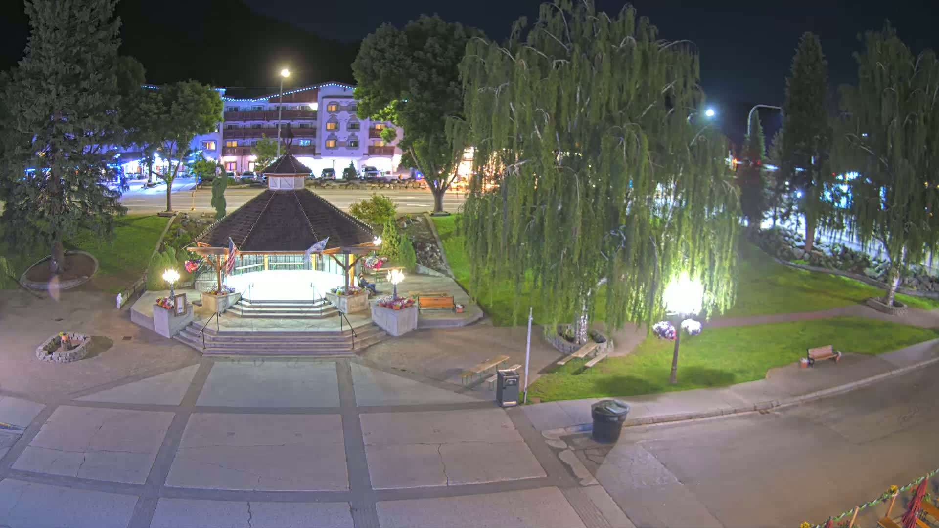 A nighttime view of a town square featuring a gazebo, lush greenery, and a hotel in the background under clear skies.