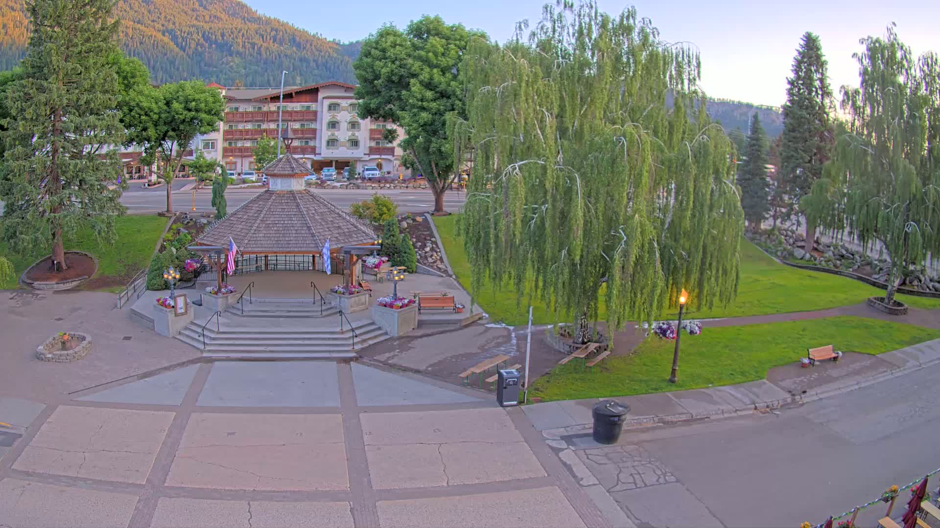 A gazebo with steps sits in a town square, surrounded by green grass, trees, and a building in the background, under a clear sky.