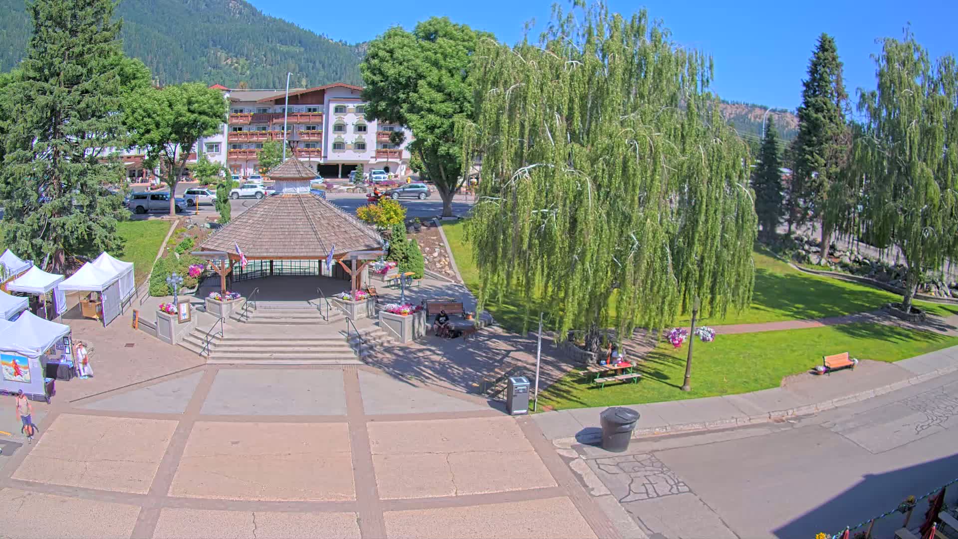 A town square with a gazebo, several trees, and vendor tents sits under a sunny sky, backed by a mountain range and a multi-story building.