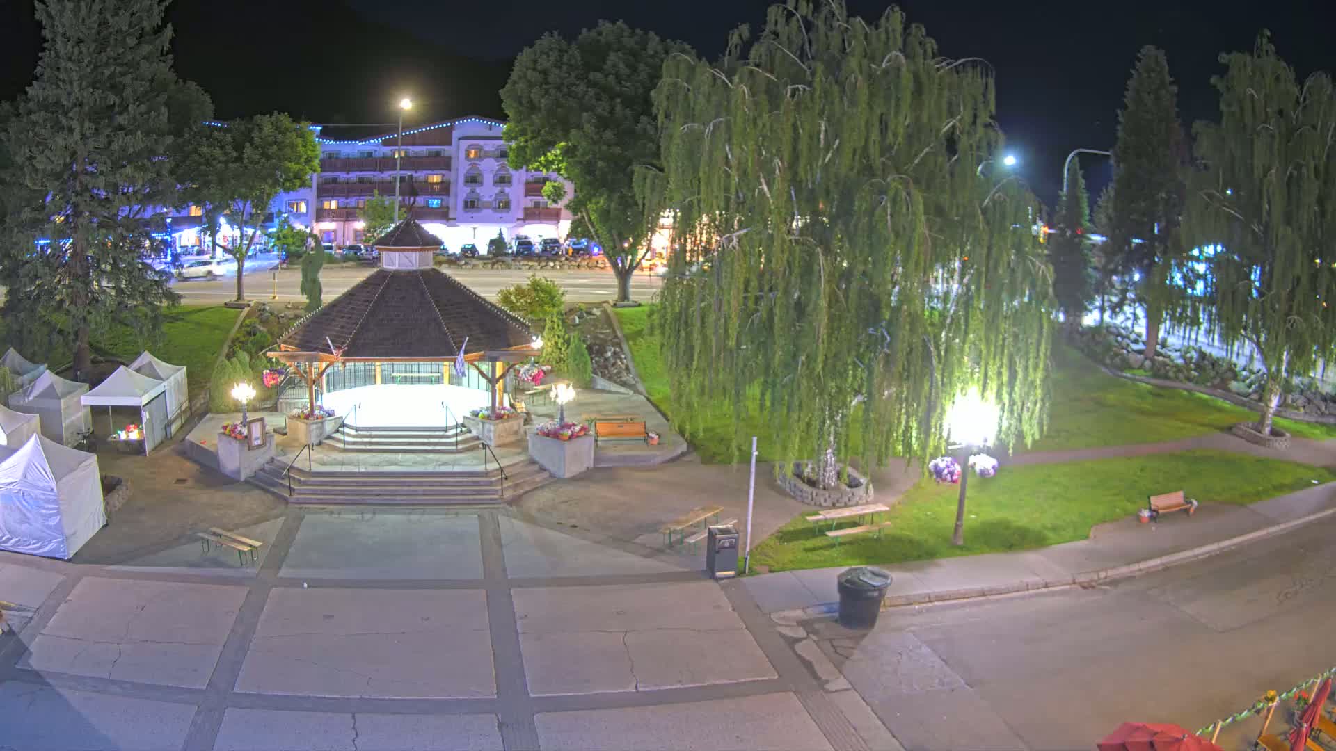 A town square with a gazebo, several trees, and vendor tents sits under a sunny sky, backed by a mountain range and a multi-story building.
