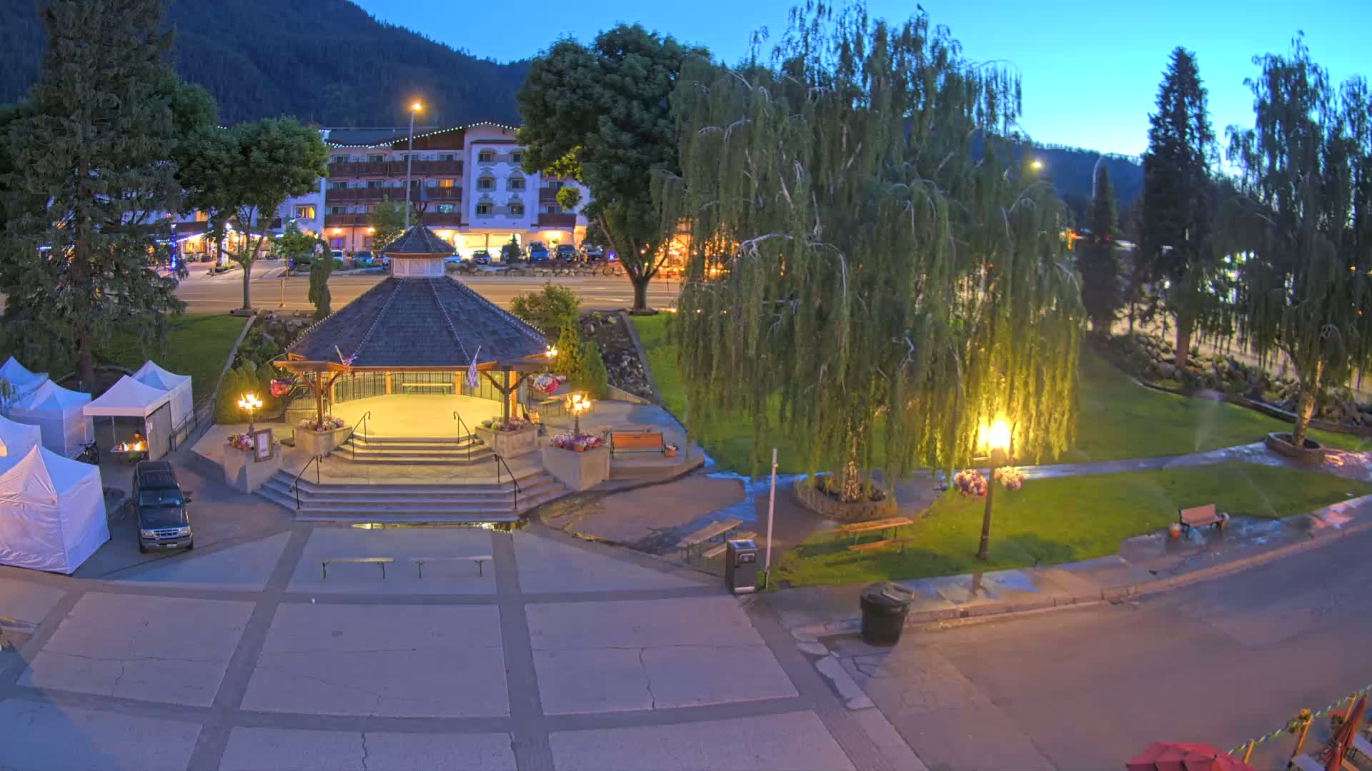 A town square at dusk, featuring a gazebo, a grassy area with trees and landscaping, and a paved area with several white tents and a vehicle parked nearby.