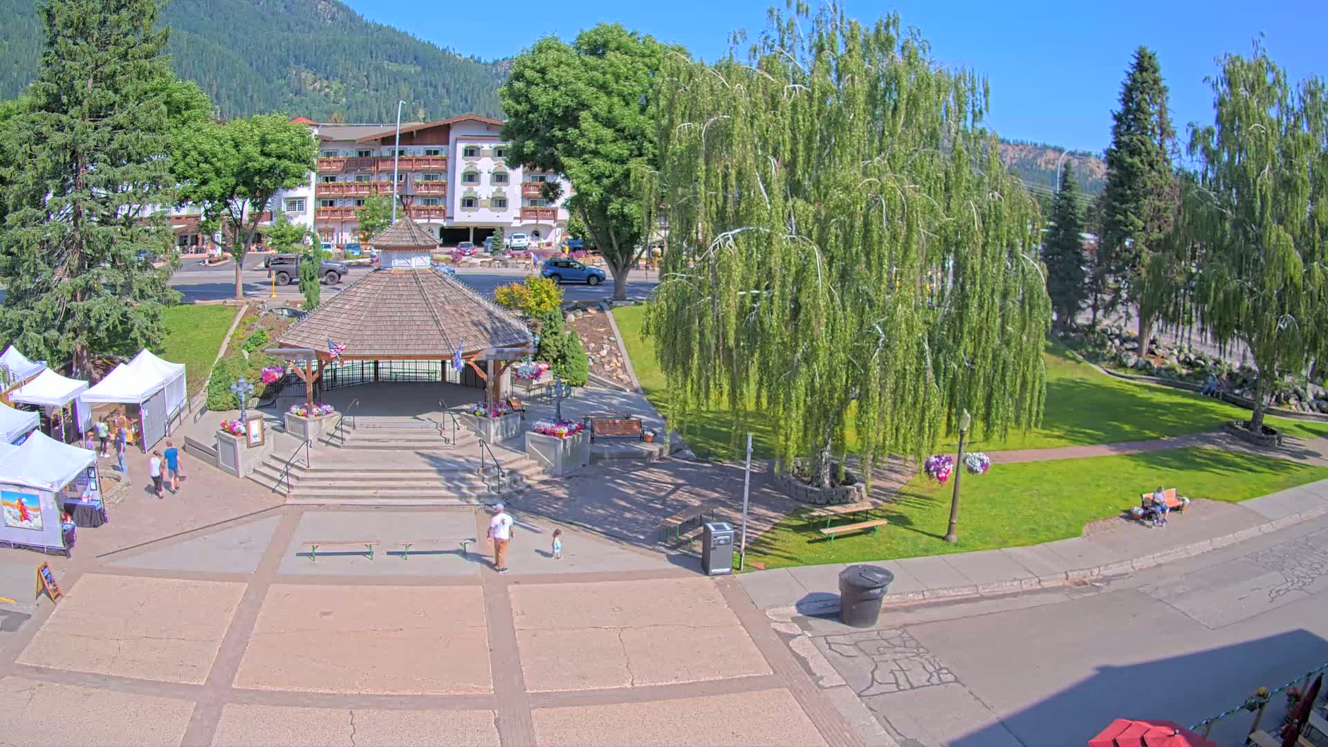 A town square with a gazebo, surrounded by green grass, trees, and a few people, under a clear sunny sky, features a small art market and a hotel in the background.